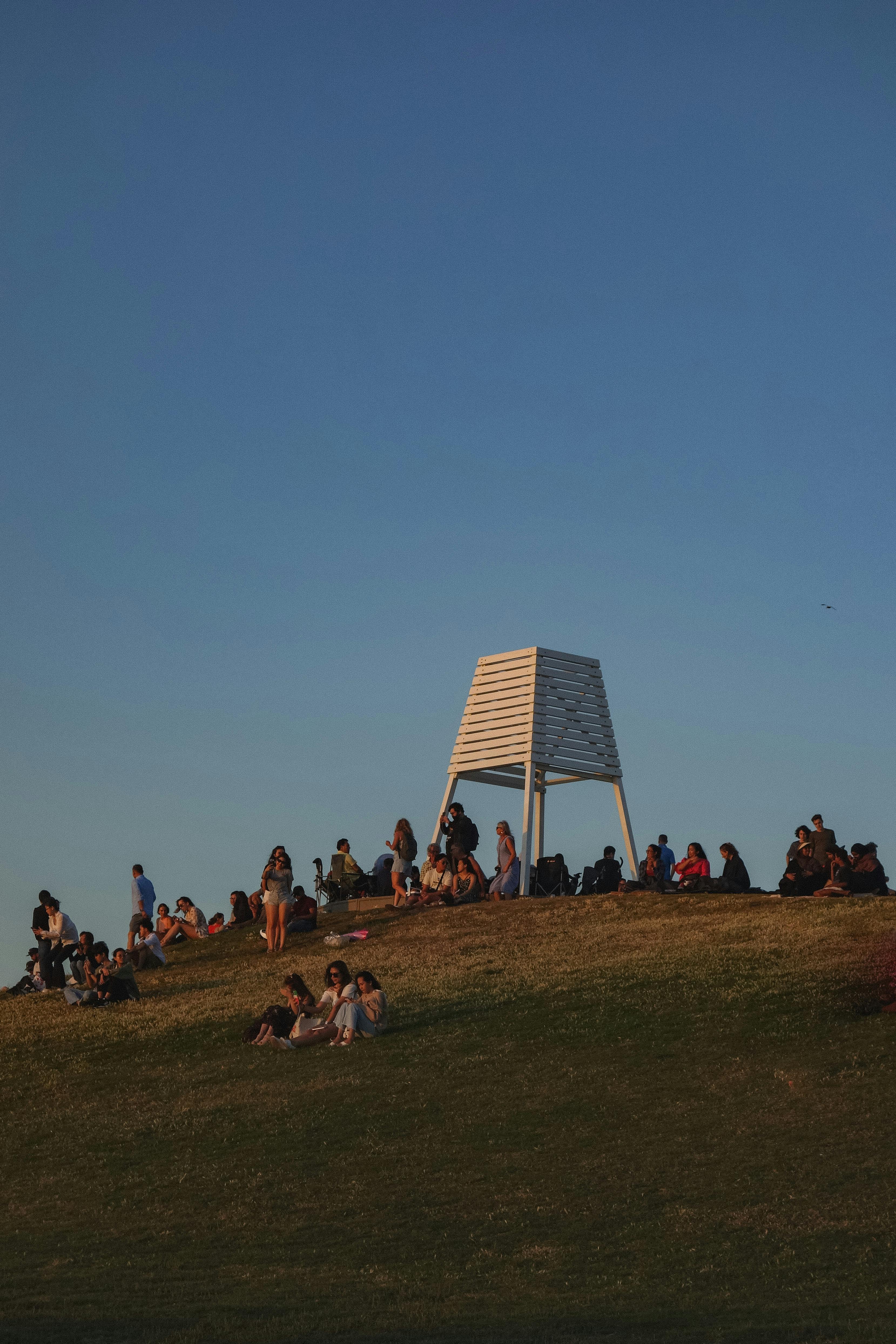Gratis Un gruppo di persone si rilassa sulla cima di una collina sotto un cielo azzurro e terso durante il tramonto, a simboleggiare un tranquillo svago all'aria aperta. Foto a disposizione