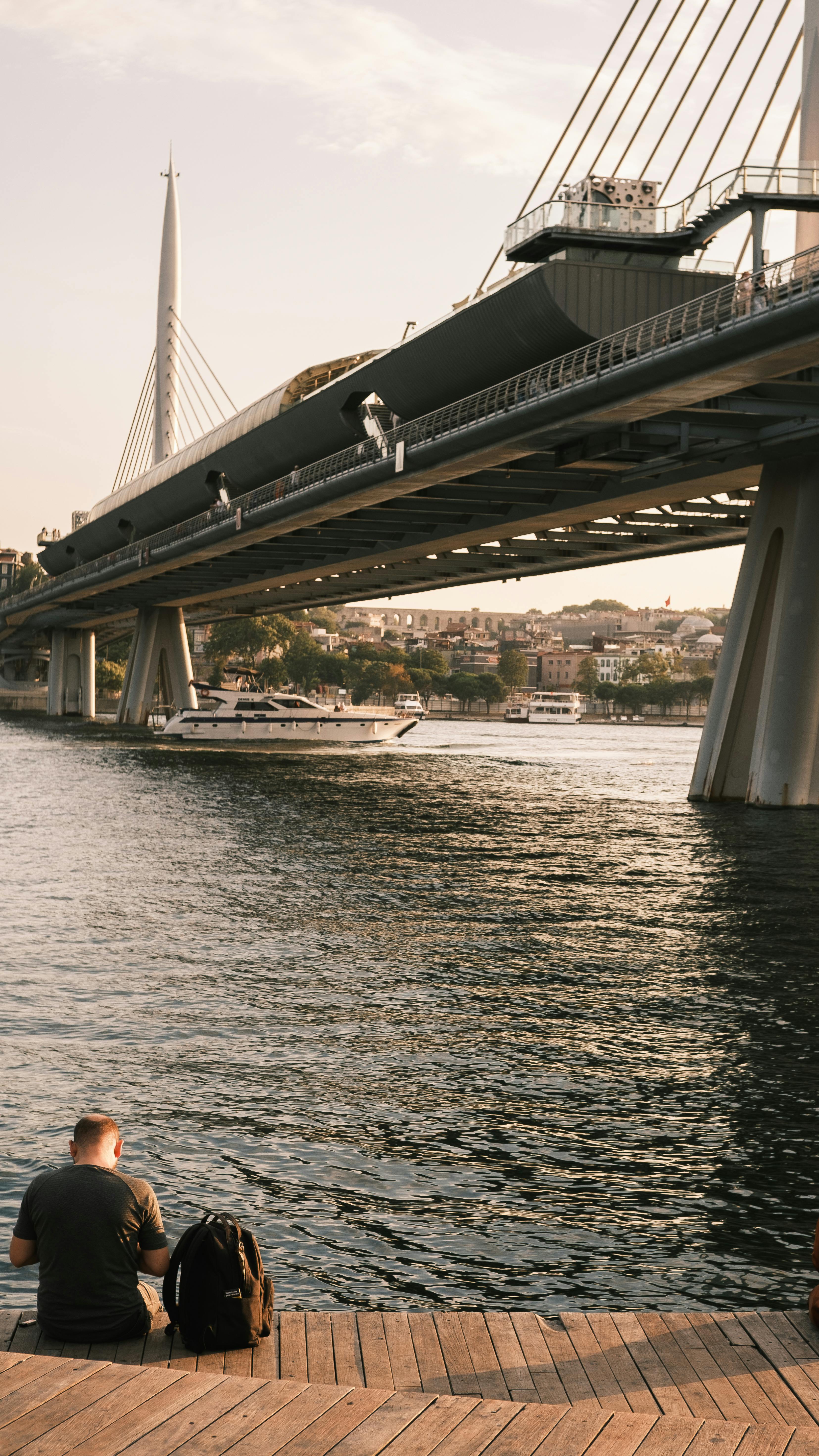 Gratuit Un homme assis au bord de l'eau, avec vue sur l'emblématique pont du métro de la Corne d'Or au coucher du soleil. Photos