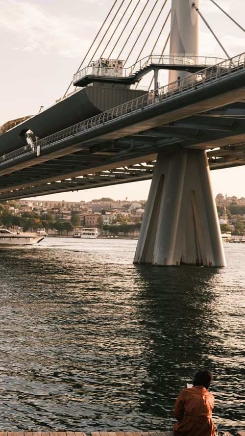 Free A person sitting by a river under a suspension bridge at sunset with urban scenery in the background. Stock Photo