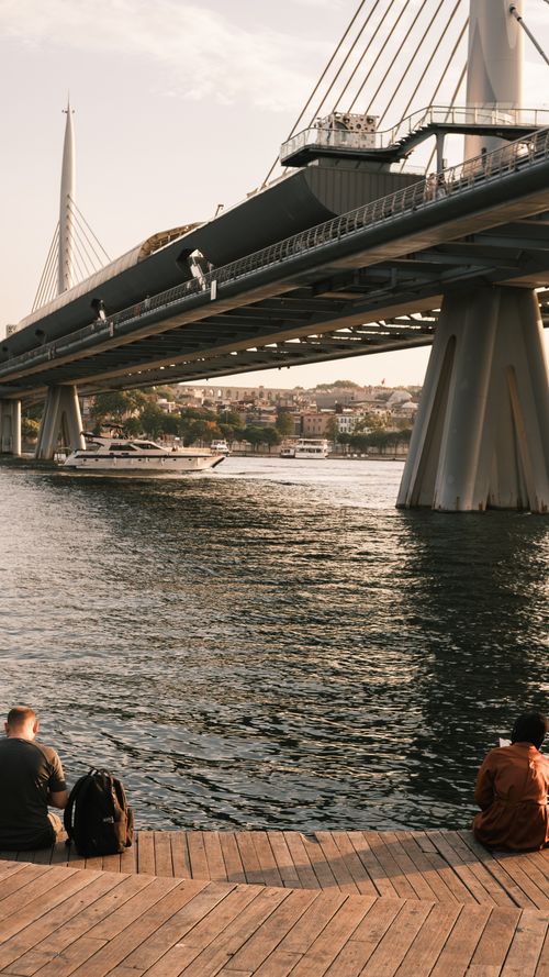 Free Two people relaxing by a river with a modern bridge in the background on a sunny day. Stock Photo