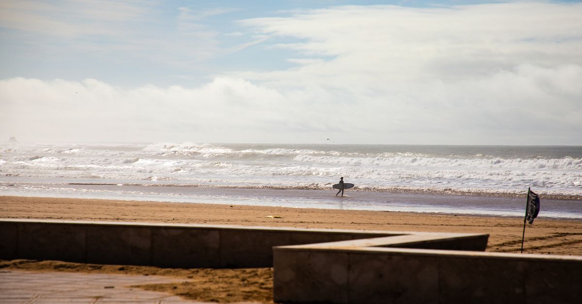 A lone surfer walks along a sunny Moroccan beach with waves and blue sky in the background.