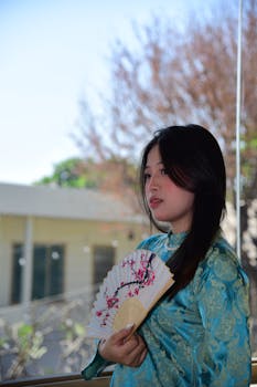 Young woman in traditional blue dress holding a decorative fan near a window.