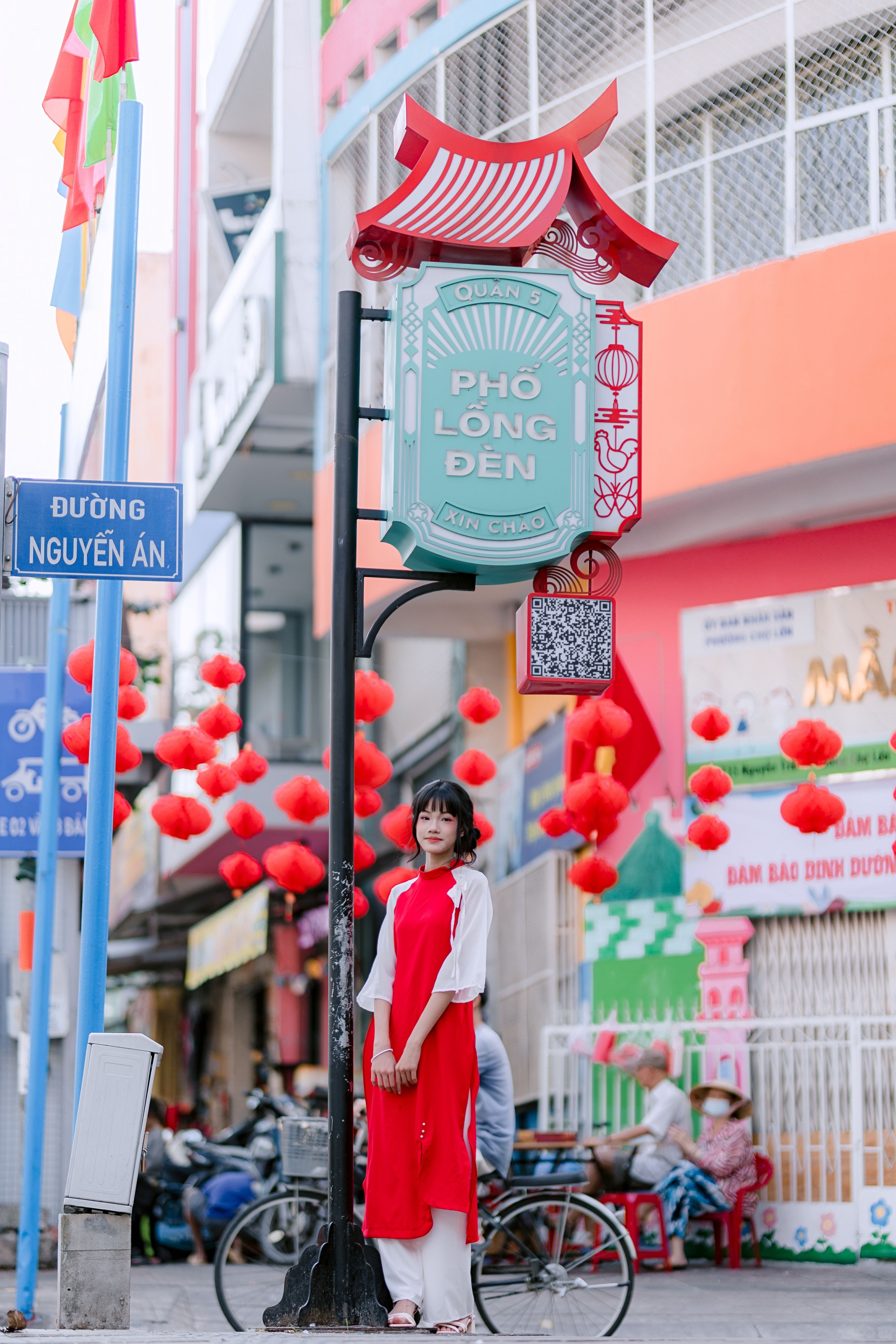 A woman in a red traditional dress poses on a vibrant street decorated with red lanterns.