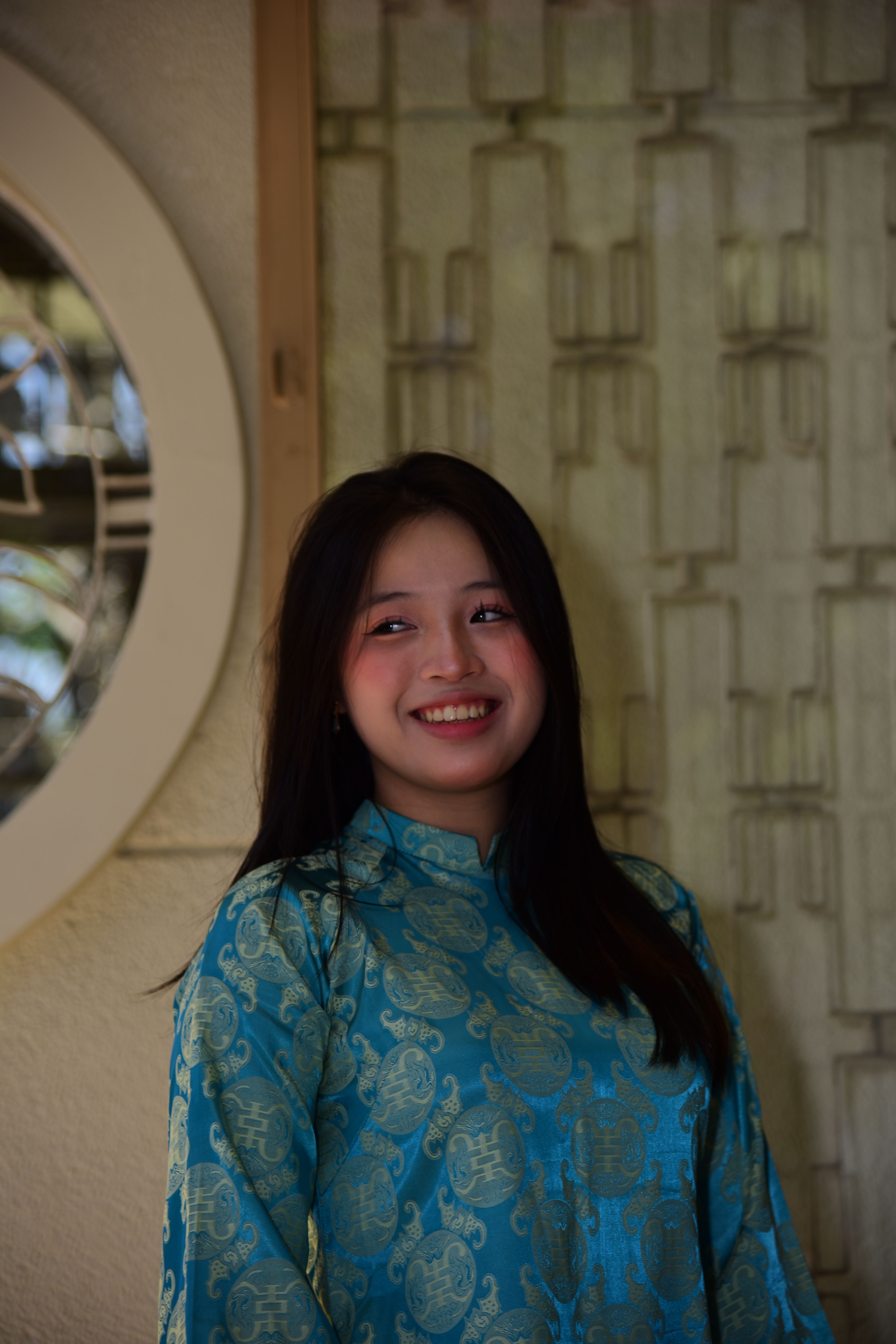 Young woman in blue traditional clothing smiling indoors against decorative background.