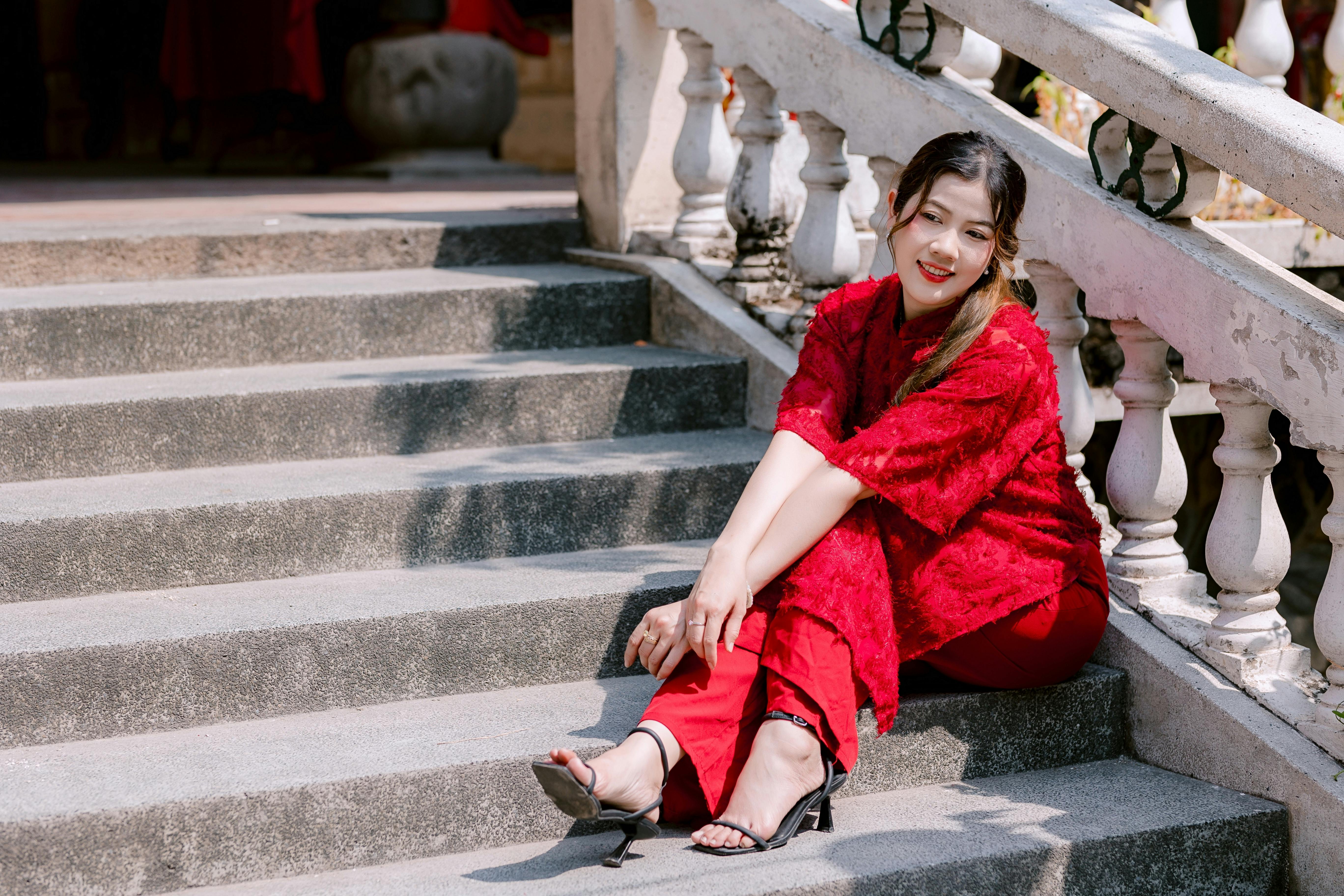 Ücretsiz Kırmızı elbiseli bir kadın, gün ışığında dış merdivenlerde oturmuş gülümsüyor. Stok Fotoğraflar