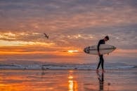 日落时分，一名冲浪者携冲浪板走在亨廷顿海滩潮湿的沙滩上，景象令人惊叹