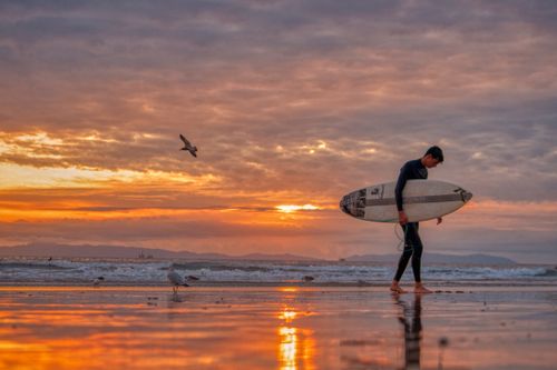 Free A surfer carrying a surfboard on Huntington Beach during a stunning sunset, reflecting on wet sand. Stock Photo