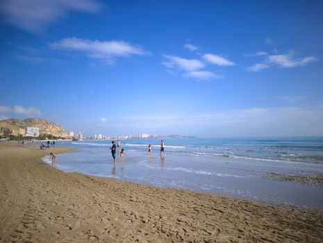 A scenic view of a family playing on the beach in Alicante, Spain under a clear blue sky.