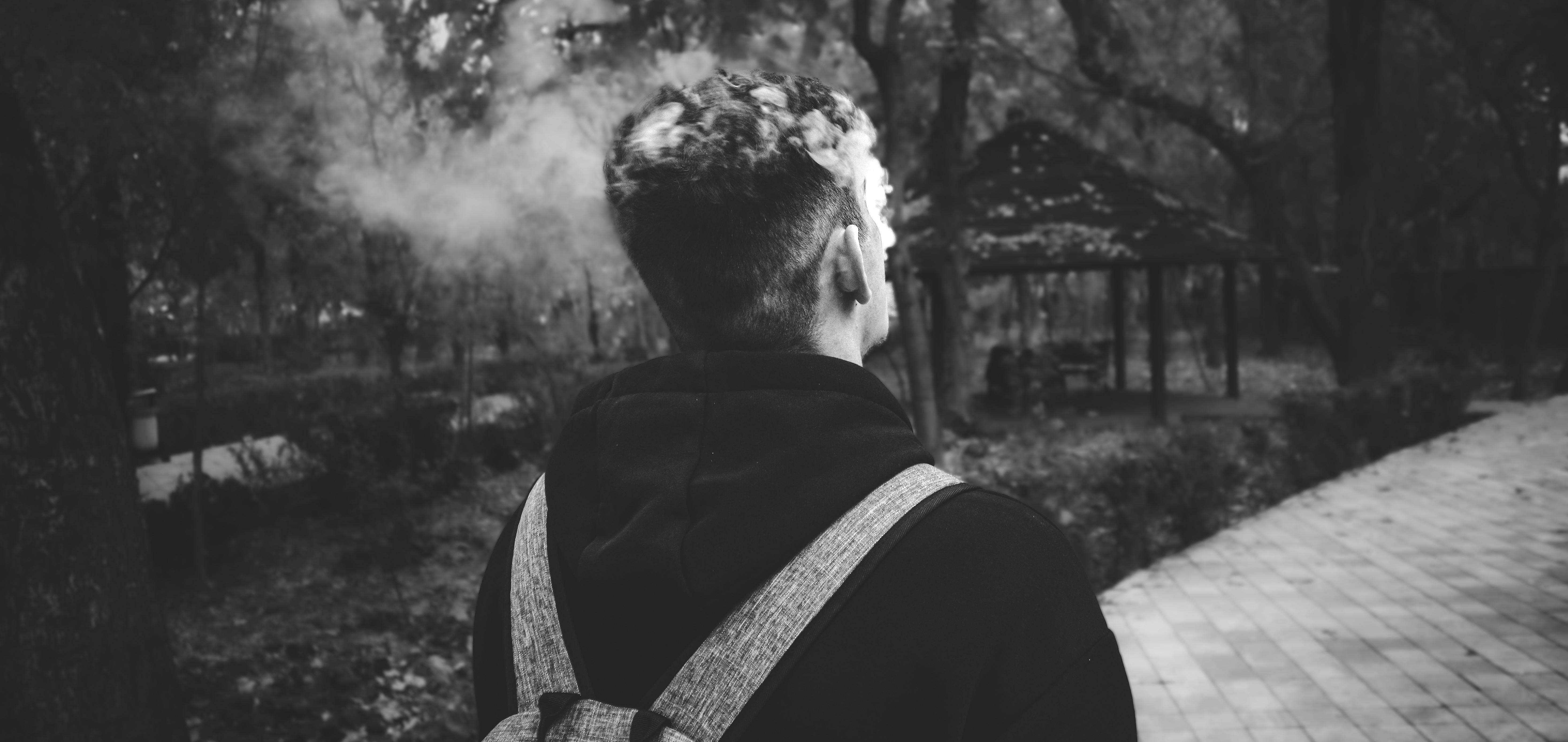 Kostenlos Ein Schwarzweißfoto einer Person, die in einem Park spaziert und Rauch ausatmet. Stock-Foto