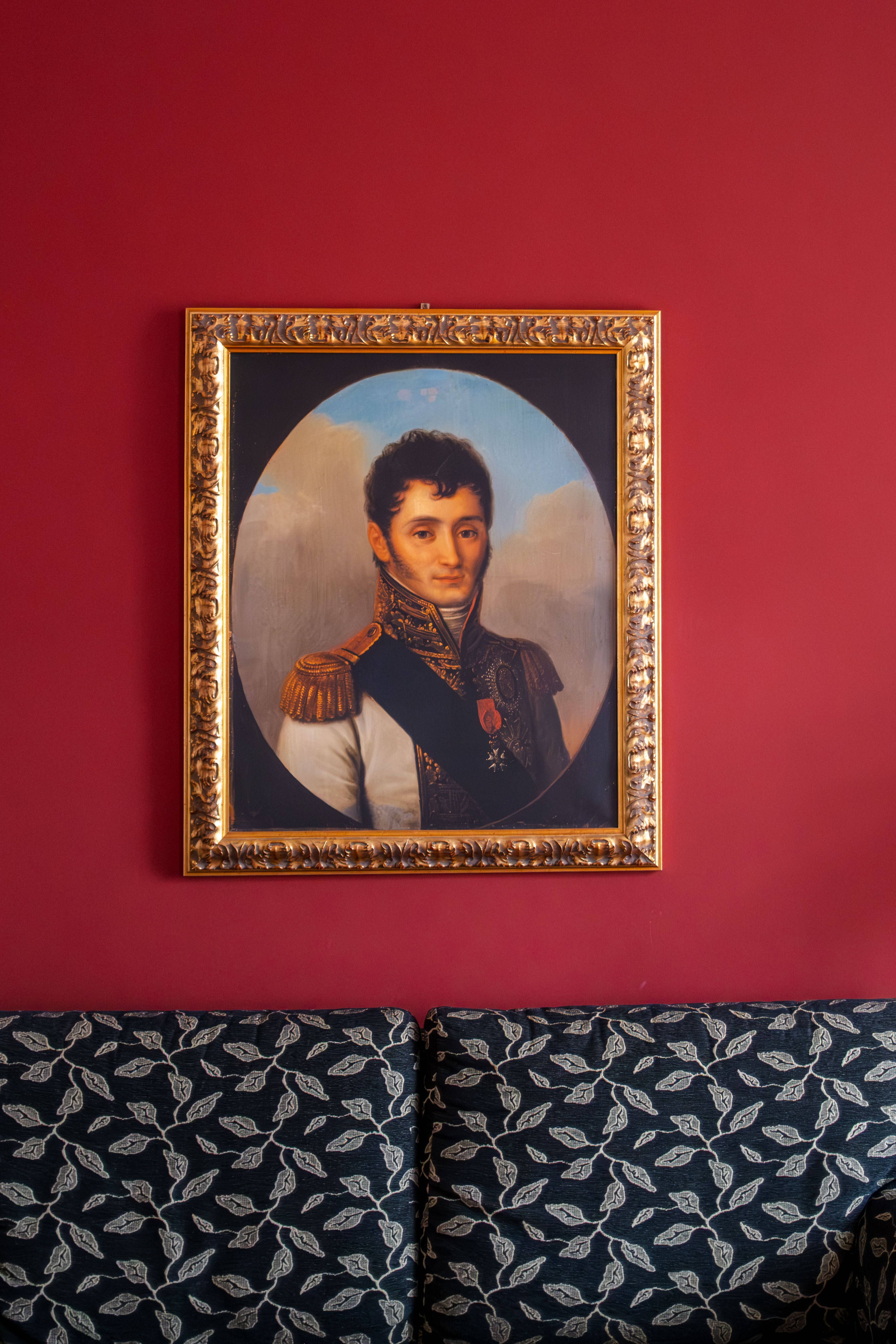 A framed historical portrait on a red wall above a patterned couch.