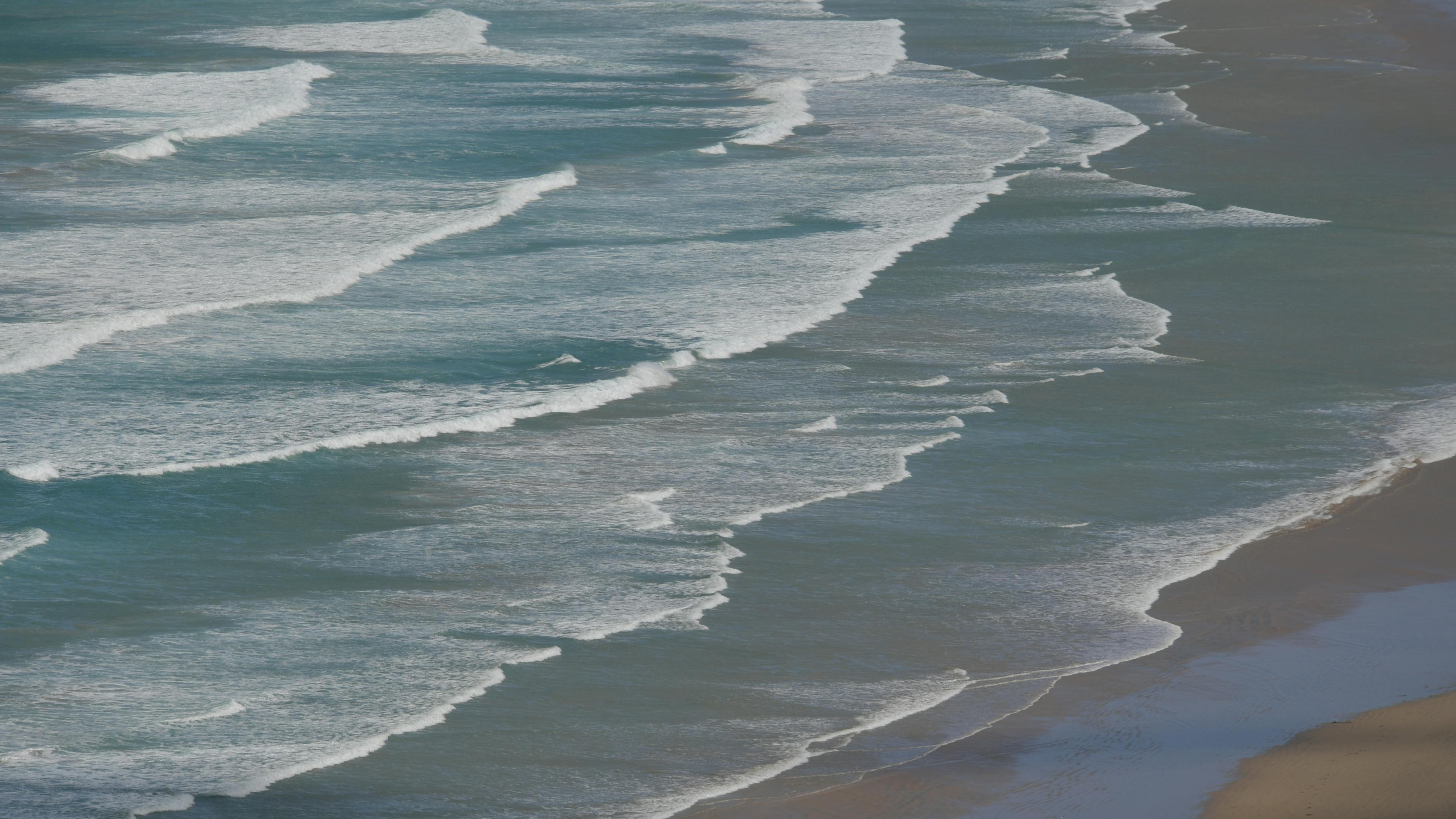 grátis Vista aérea de ondas oceânicas quebrando dinamicamente em uma costa arenosa, com destaque para a textura da água. Foto profissional
