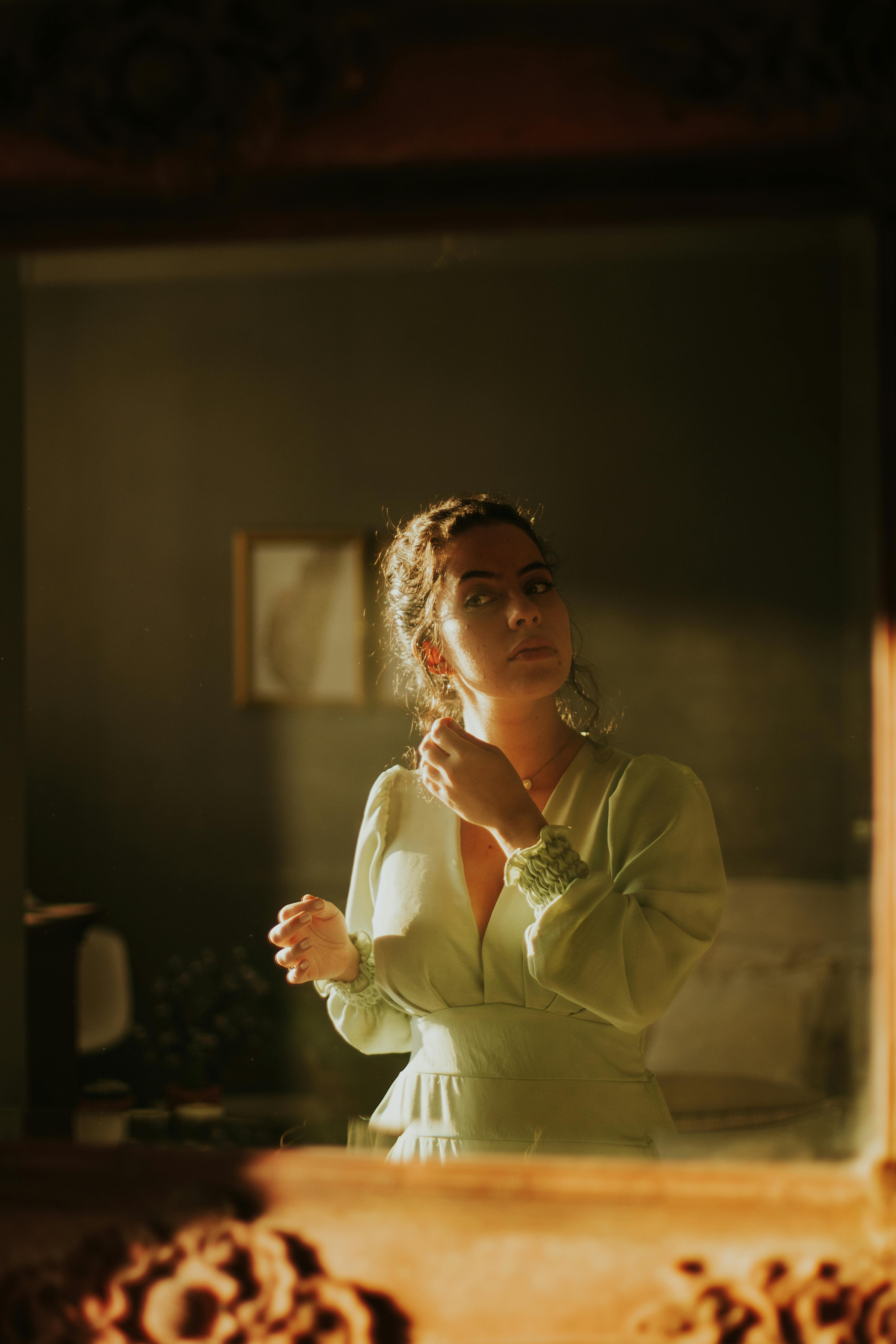 Gratis Una donna con un abito verde posa con grazia davanti a uno specchio, illuminata dolcemente dalla calda luce del sole. Foto a disposizione
