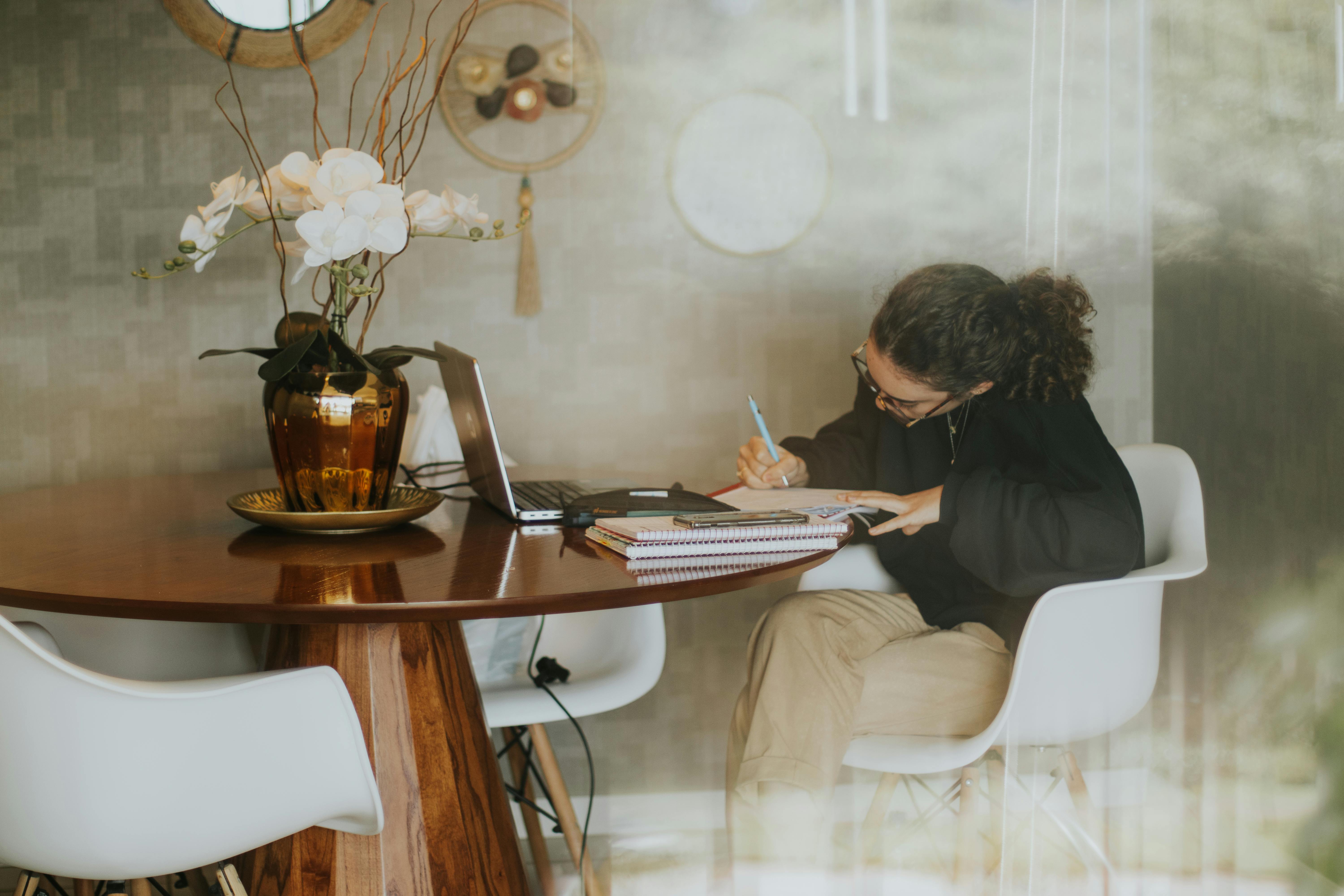 Woman writing at a table with a laptop and flower vase, creating a cozy home office ambiance.