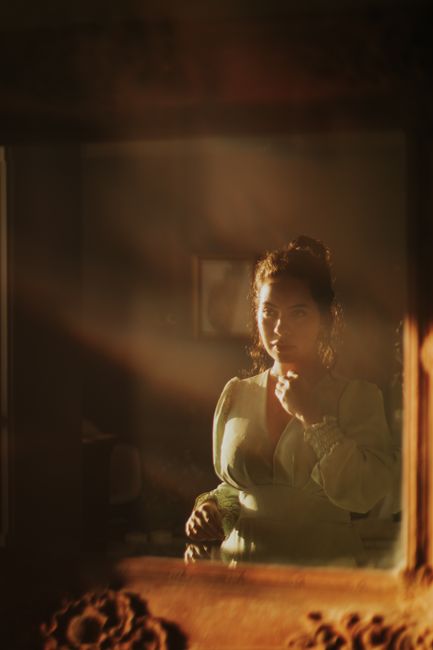 A woman in vintage attire is beautifully reflected in a dimly lit mirror, creating a moody, artistic atmosphere.