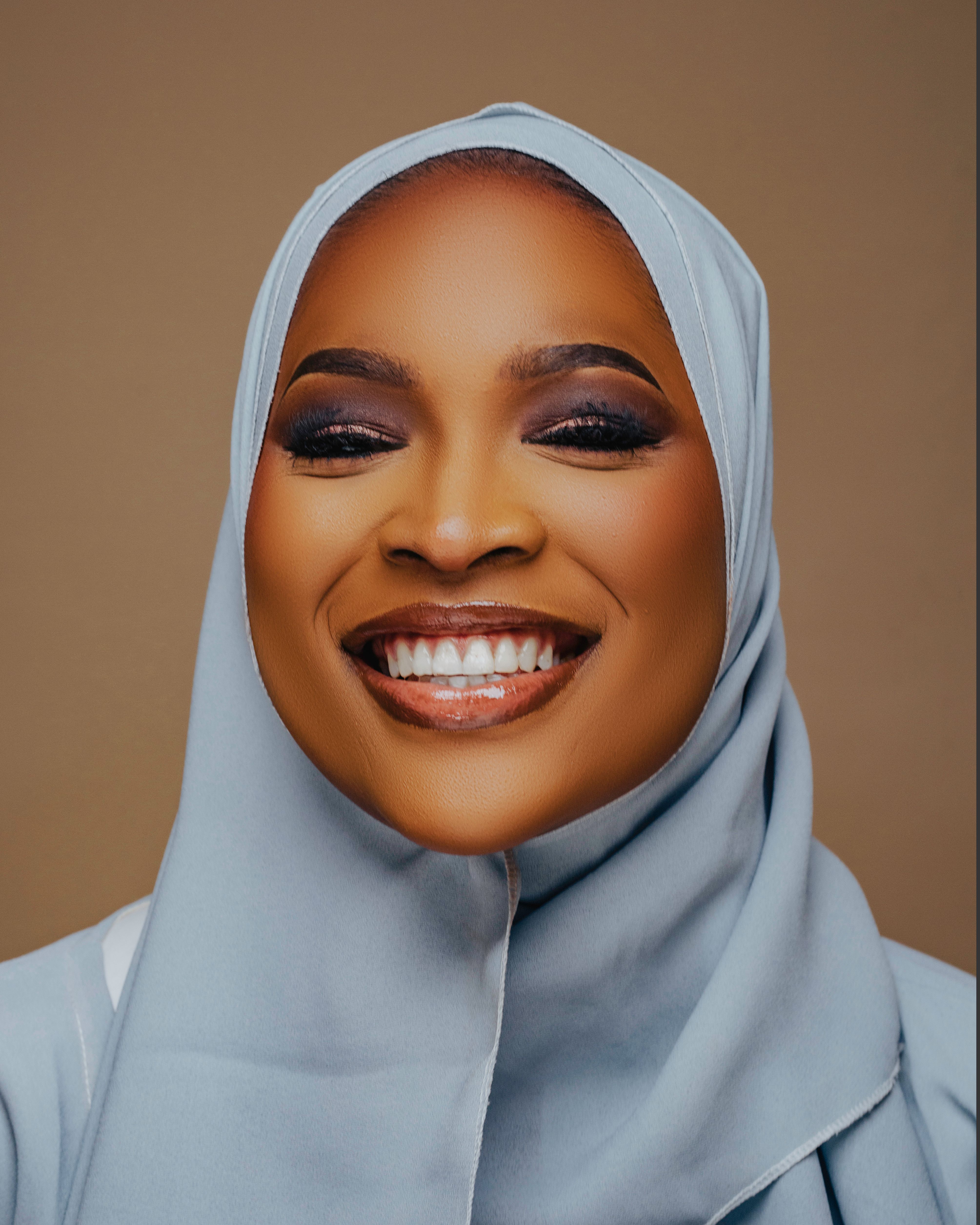 Close-up portrait of a smiling woman in a hijab with a beige background.