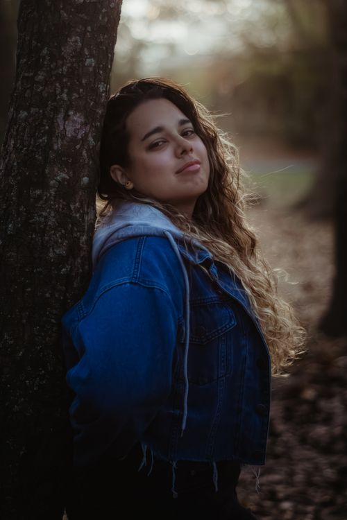 Free Portrait of a young woman with curly hair and denim jacket leaning against a tree in a serene autumn forest. Stock Photo