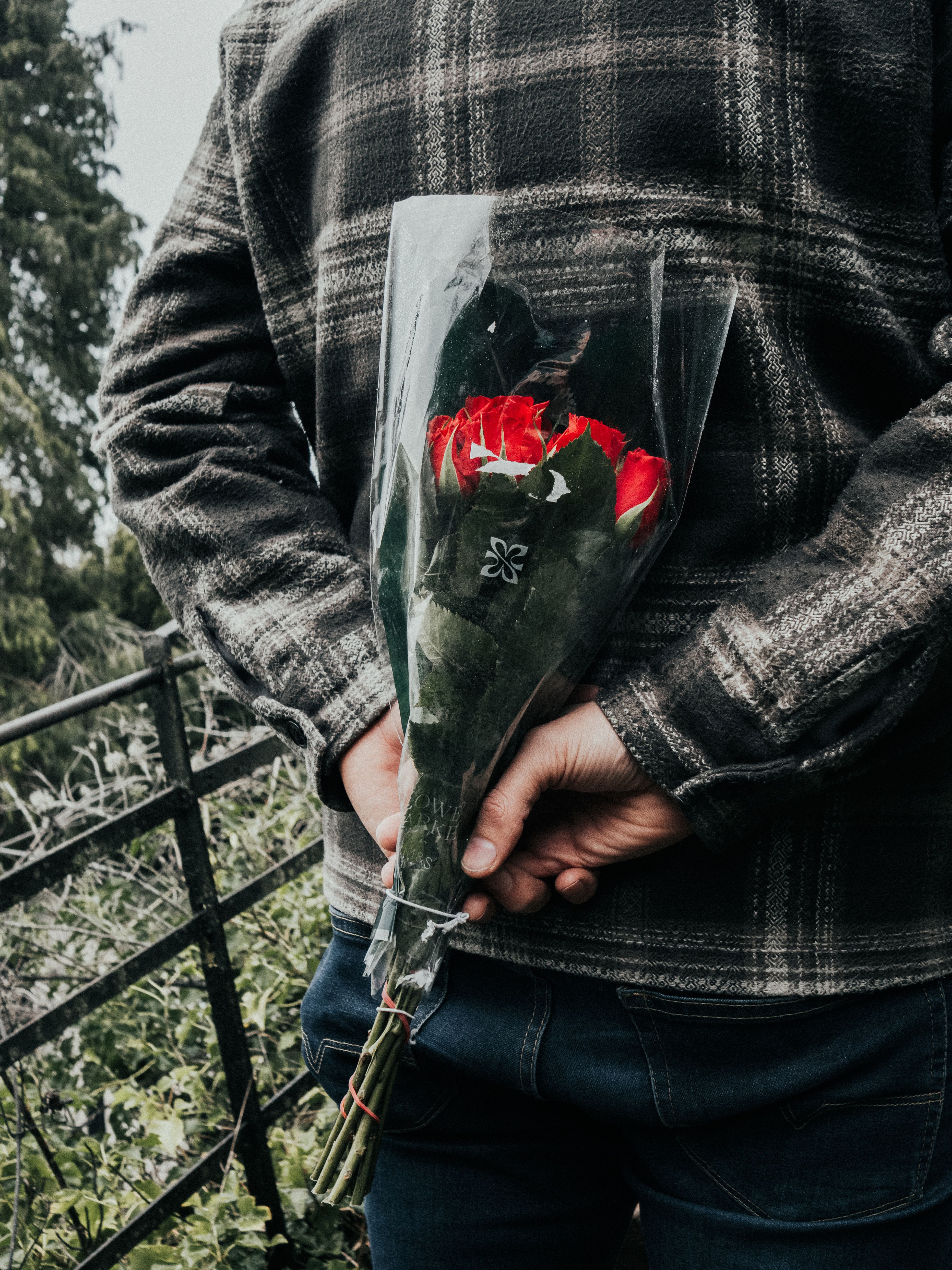 A man in a plaid jacket holds a bouquet of red roses behind his back on a balcony.