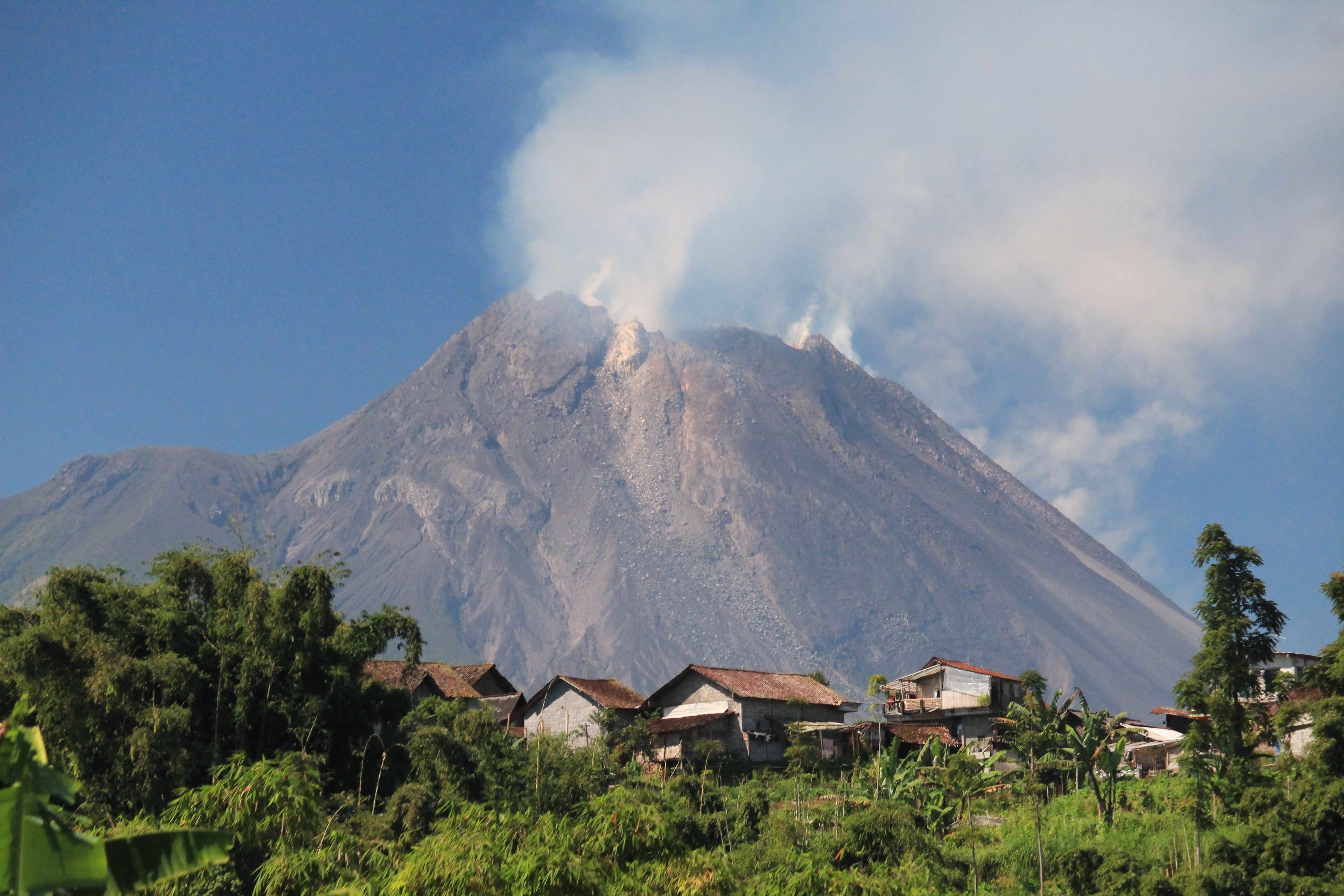 A smoking volcano looms over rooftops of a rural village under a clear blue sky.