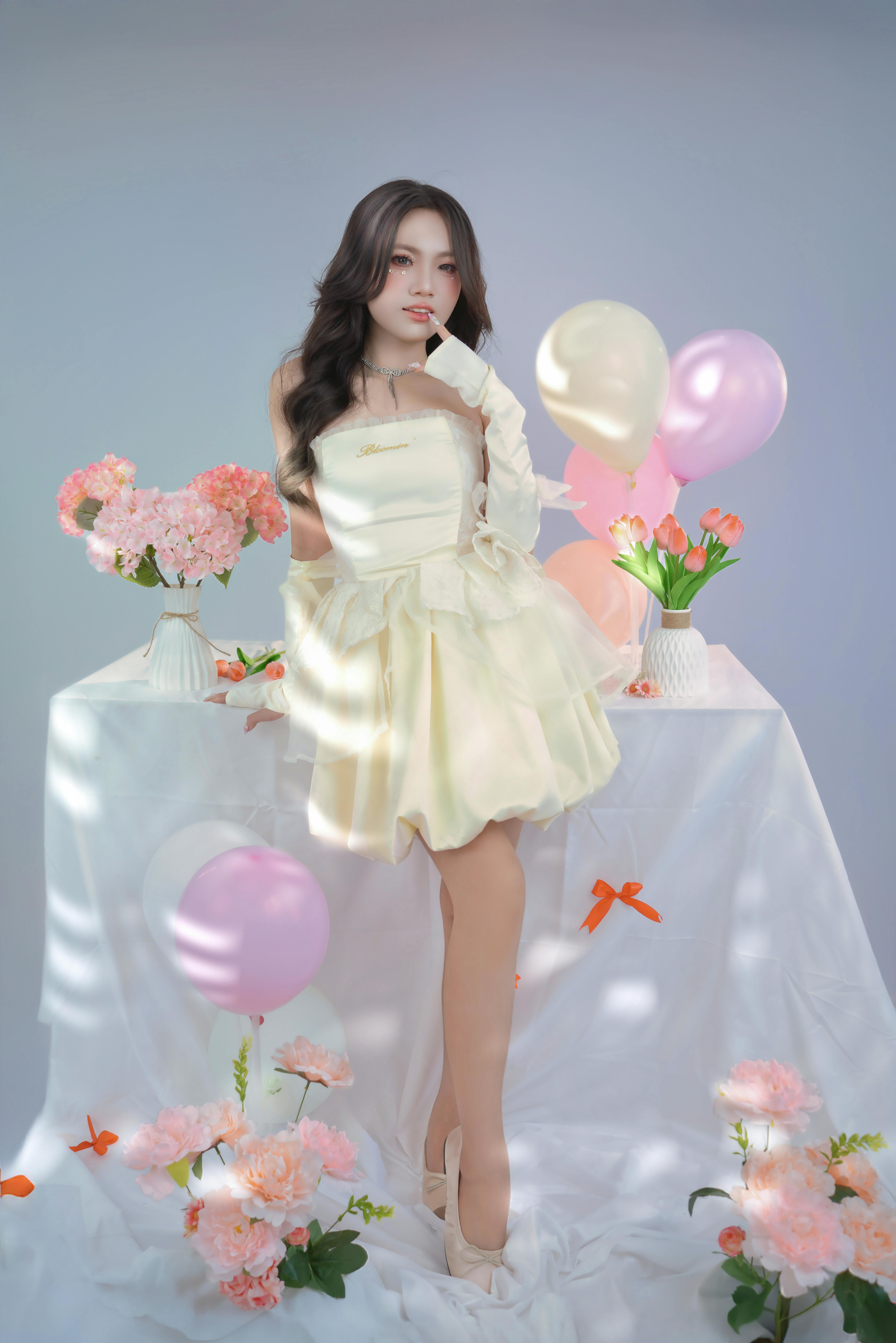 A woman in a cream dress poses elegantly with pink flowers and balloons against a soft background.