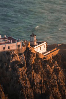 A stunning aerial view of Cap Carbon Lighthouse in Algeria bathed in warm sunset light, perched above the ocean.