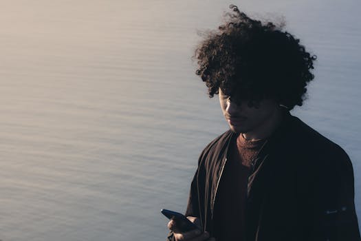 Young man with curly hair gazes at phone by the calm sea in Béjaïa, Algeria.