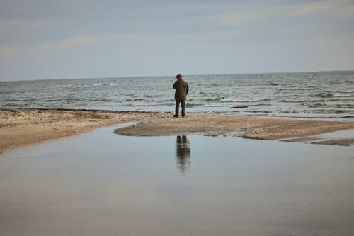 Free A lone person stands on a sandy beach, gazing at the calm sea under a cloudy sky. Stock Photo