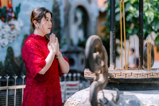 Asian woman in red dress praying at a temple with incense sticks in foreground.