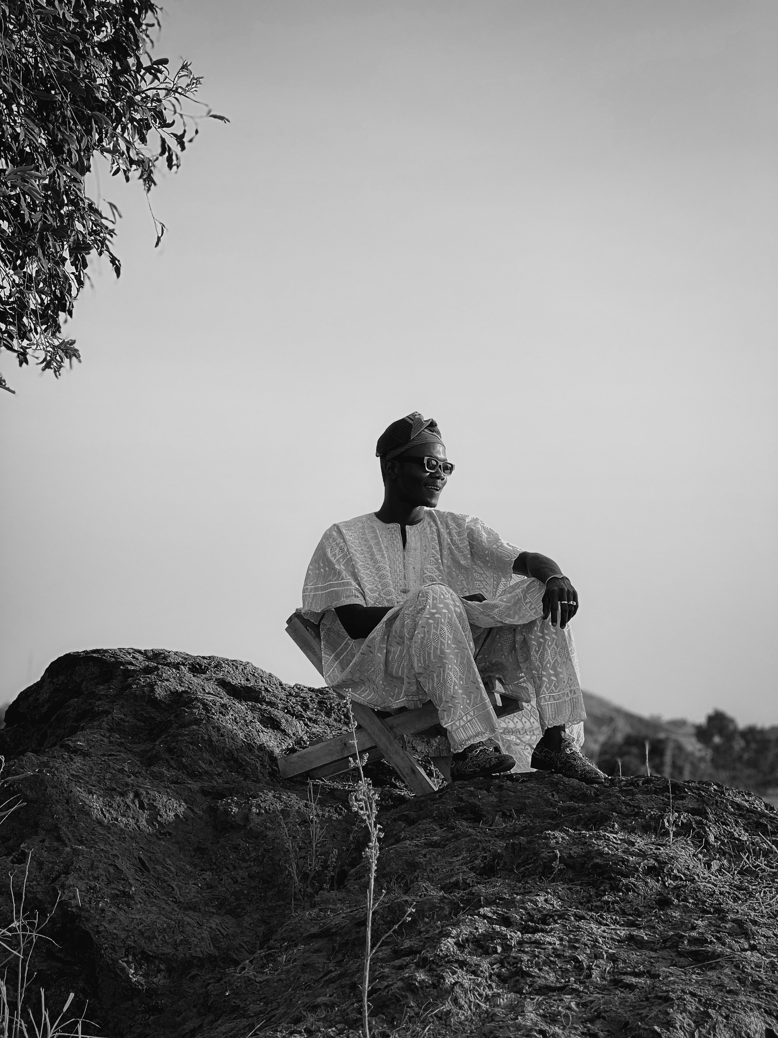 A man in traditional attire sits reflectively on a rock, surrounded by nature in black and white.