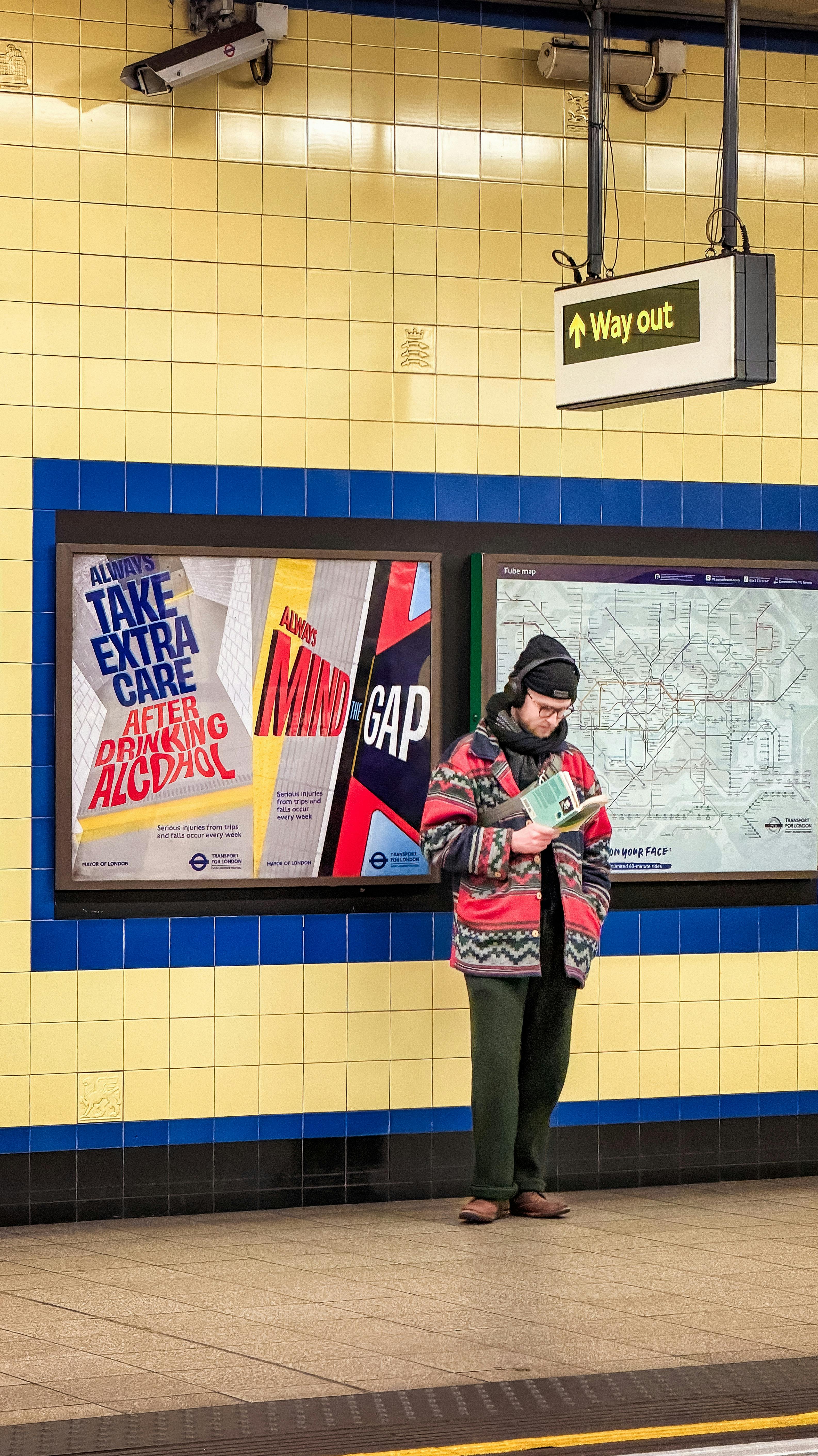 Man reading while waiting in London tube station, featuring vibrant posters and signage.