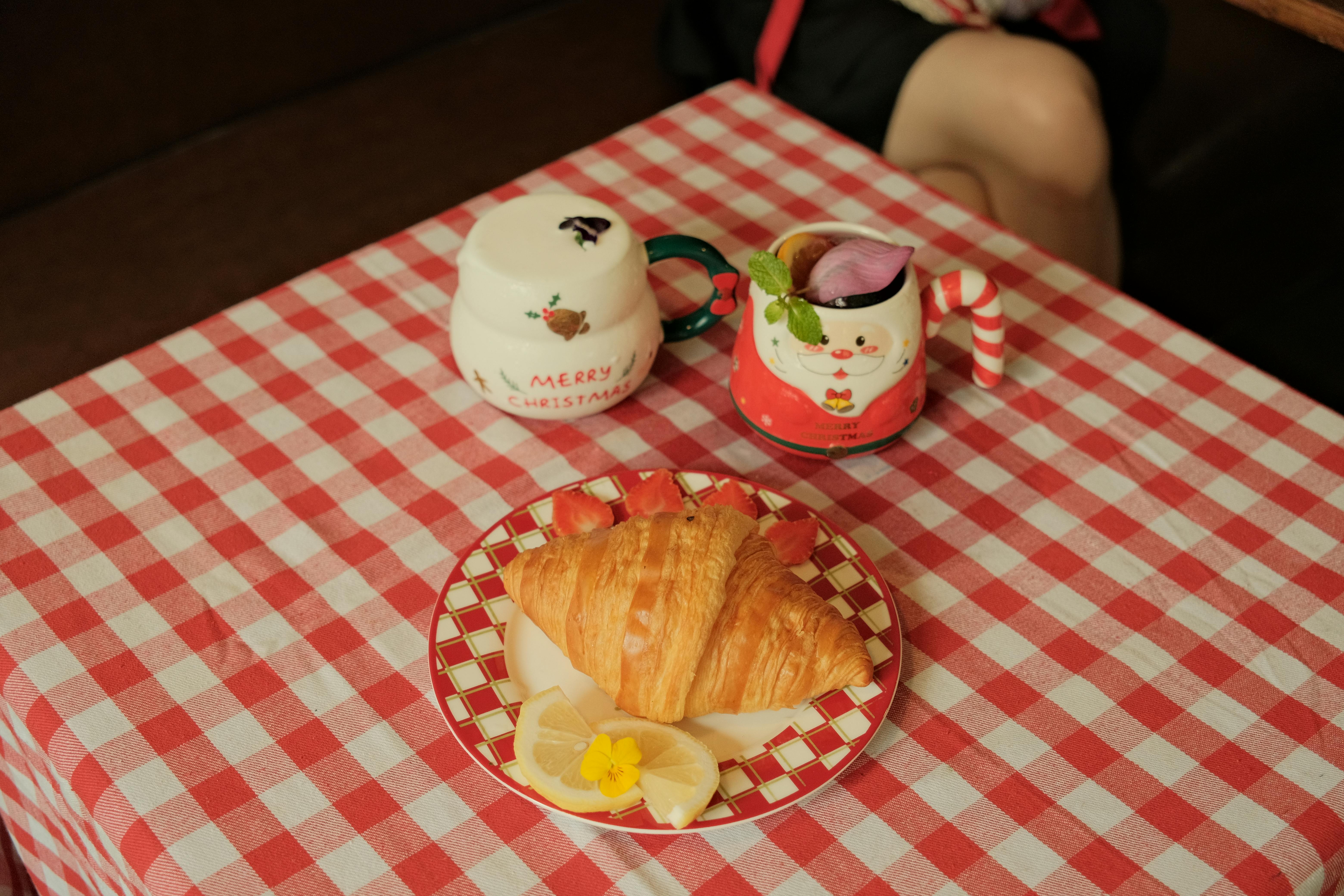 無料 赤いチェック柄のテーブルクロスの上にクロワッサンとお祝いのマグカップを並べた、楽しい朝食のセッティング。 写真素材