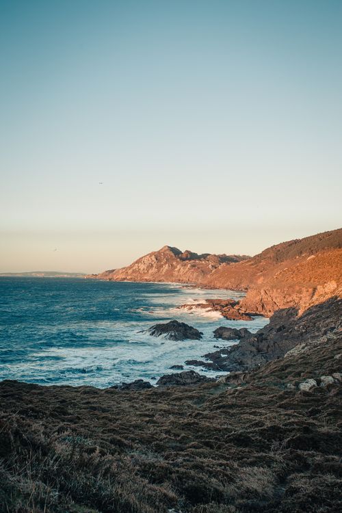 Free Spectacular view of blue ocean with foamy waves touching rough stones surrounded by mounts under cloudless sky in evening Stock Photo
