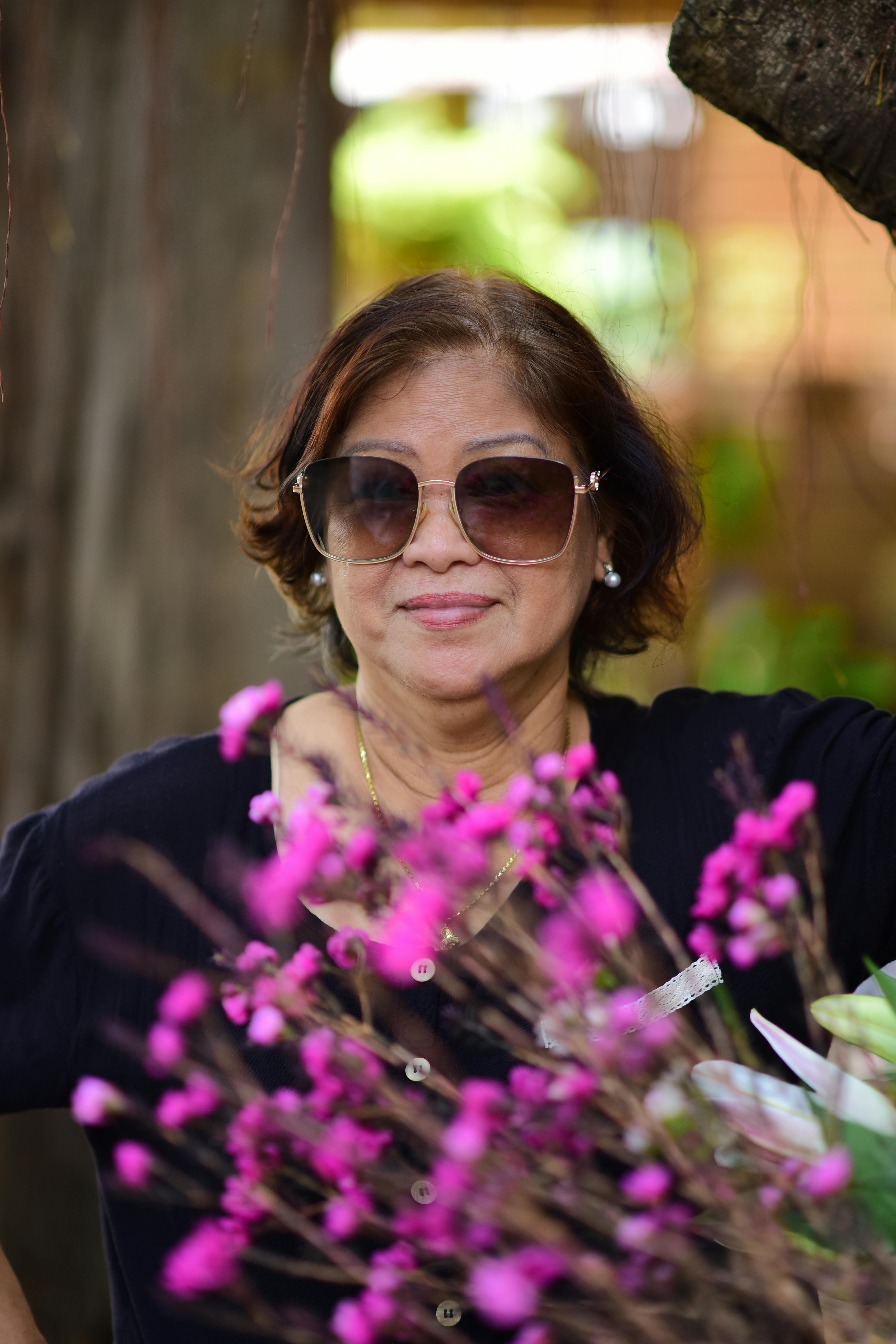Confident woman wearing sunglasses smiles beside vivid pink flowers, set against a lush outdoor background.