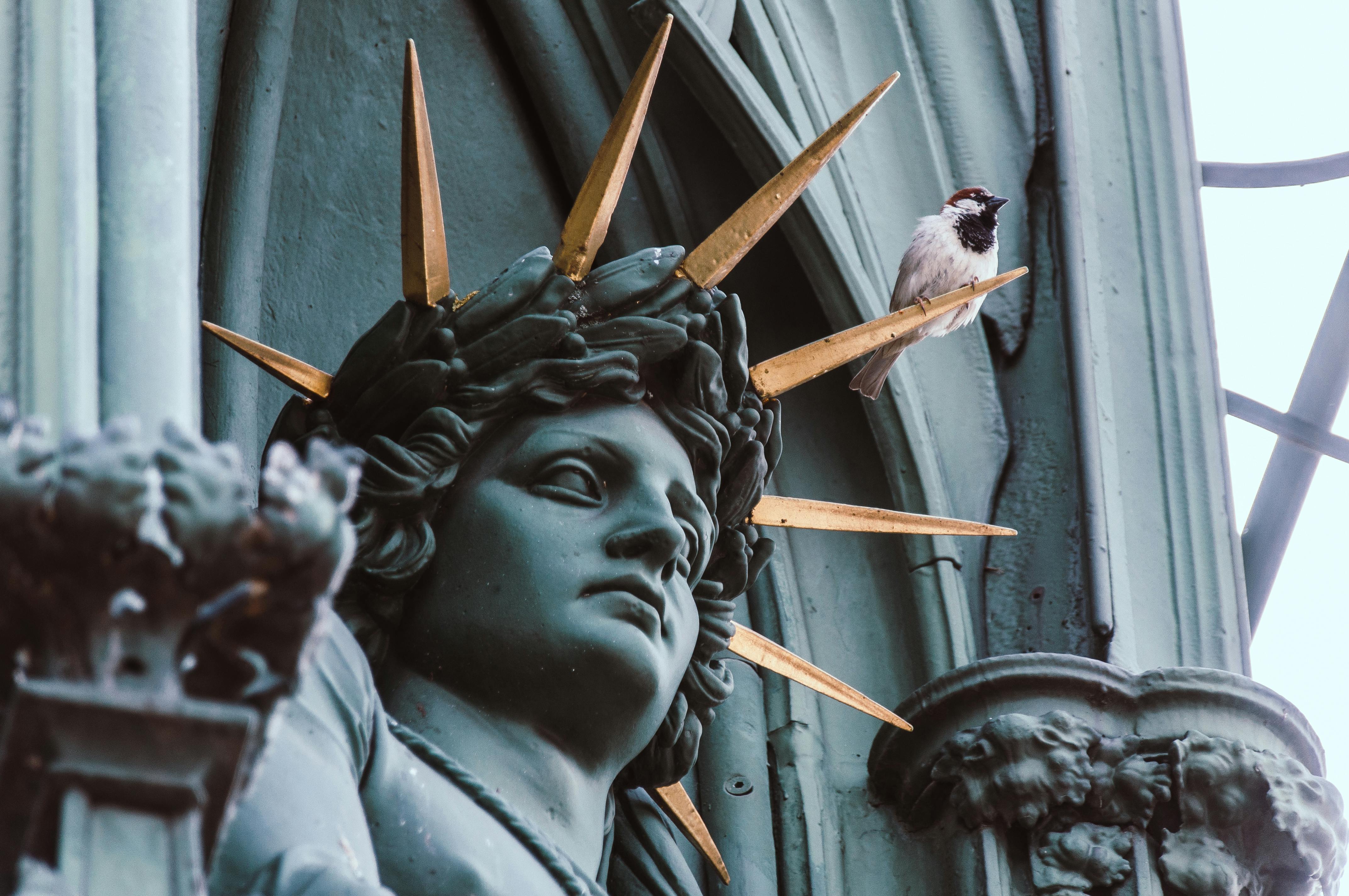 Close-up of a statue with gold spikes and a sparrow perched, symbolizing contrast between art and nature.
