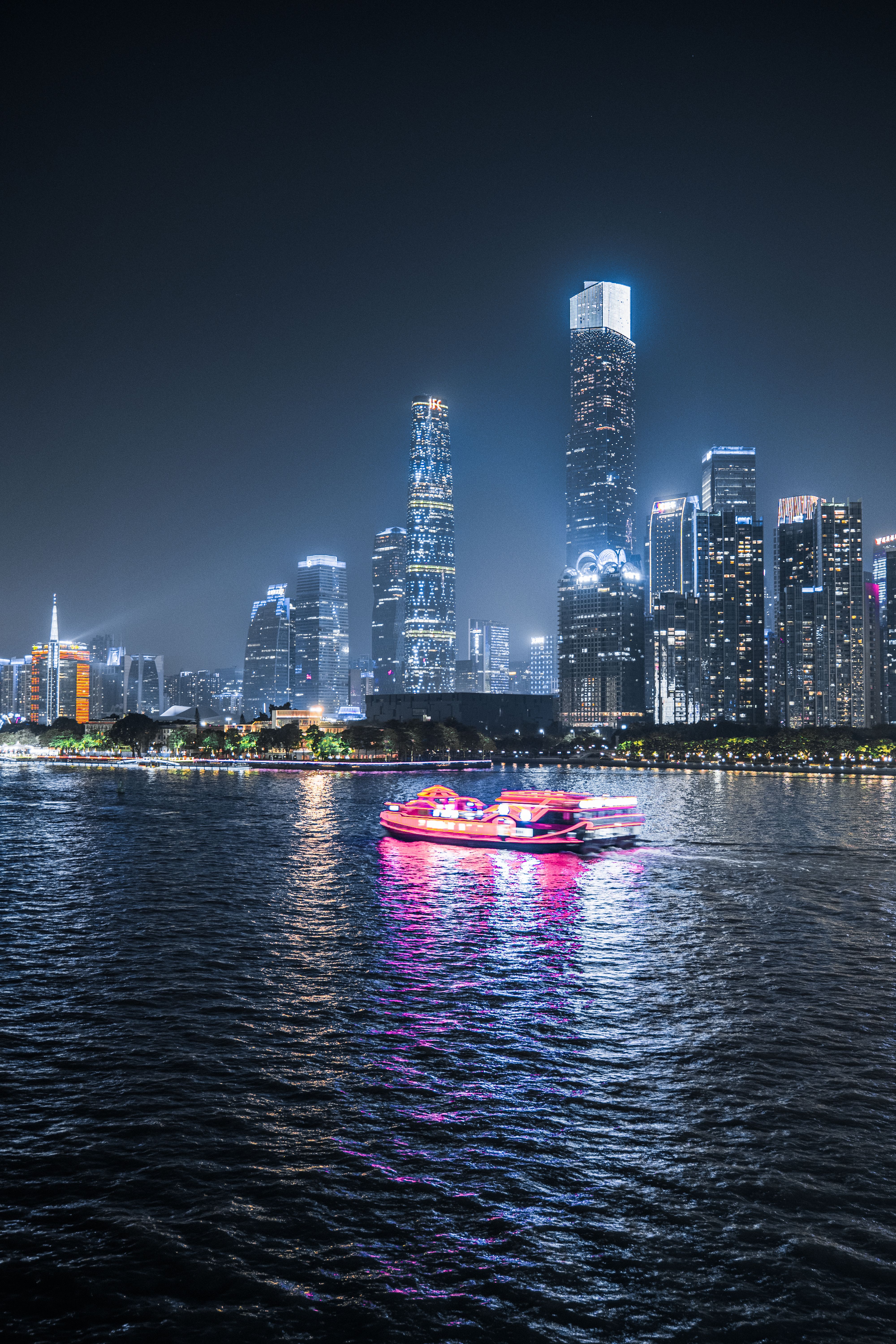 Vibrant city skyline at night with a pink-lit boat on a river reflecting lights.