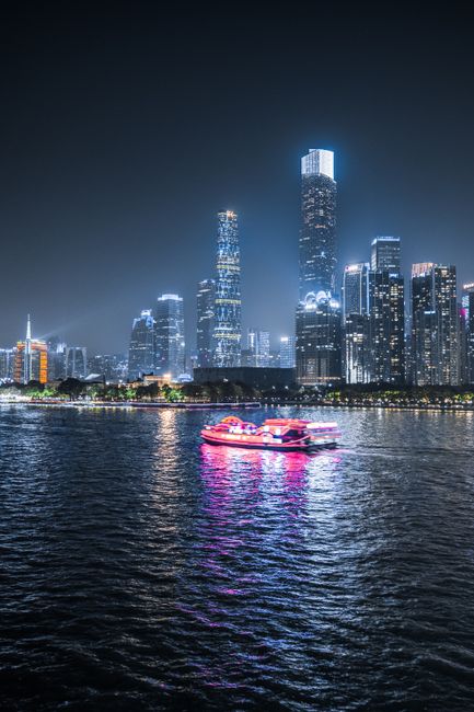 Vibrant city skyline at night with a pink-lit boat on a river reflecting lights.