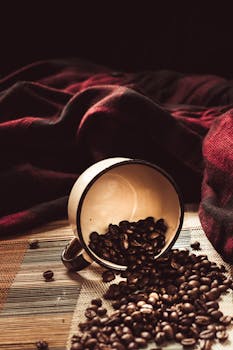 Spilled coffee beans from a rustic cup on a textured surface with a cozy blanket background.