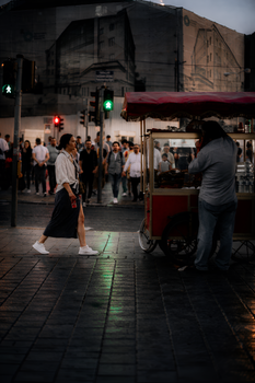 Woman walks past street vendor at evening in city, capturing urban life.