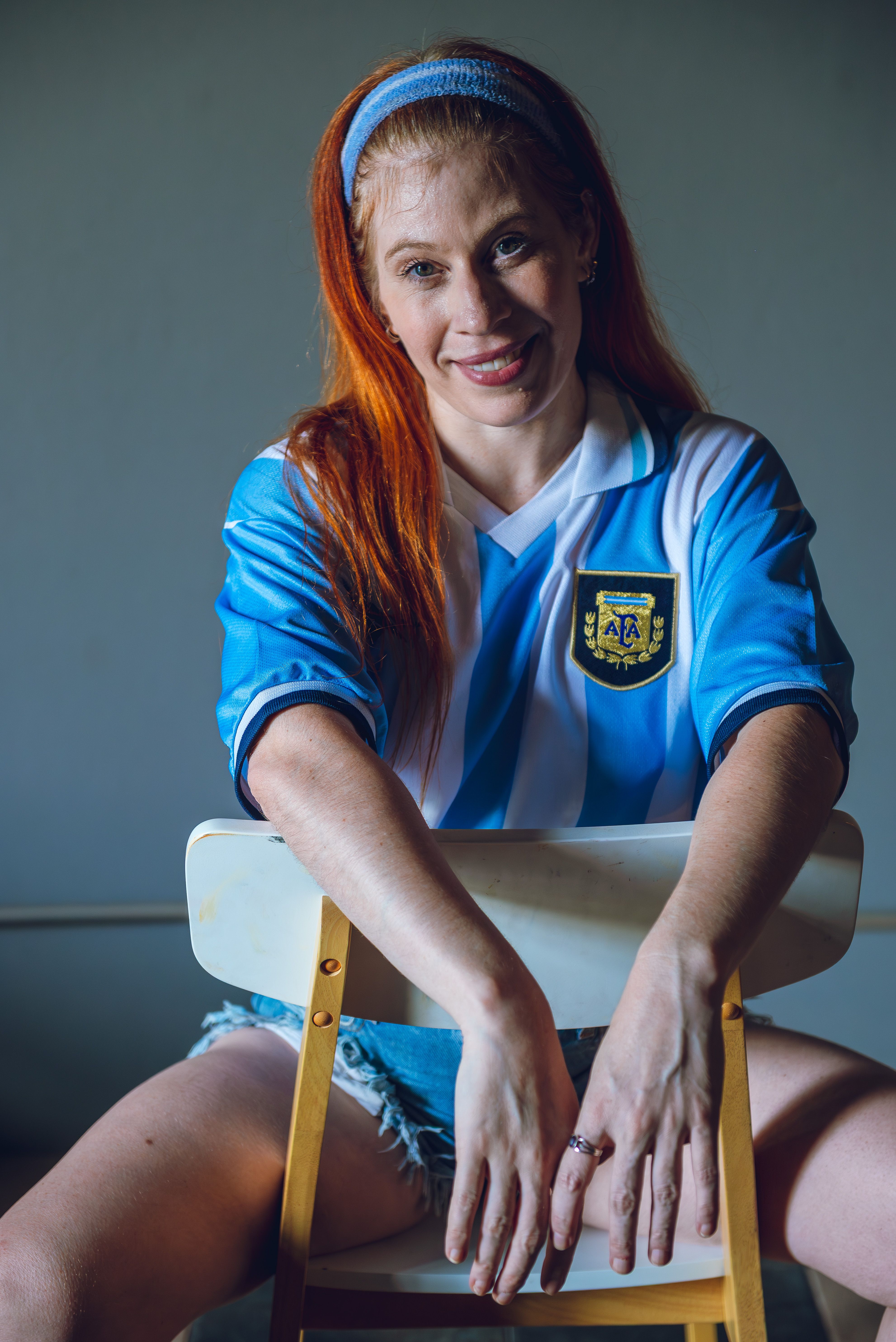Woman sitting on chair wearing Argentine soccer jersey indoors, smiling.
