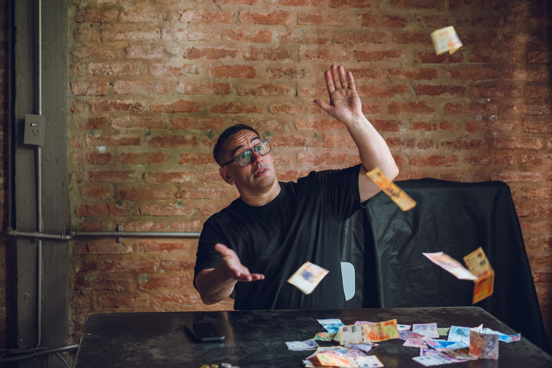 Adult man with glasses tosses Argentine Pesos joyfully in a rustic indoor setting.