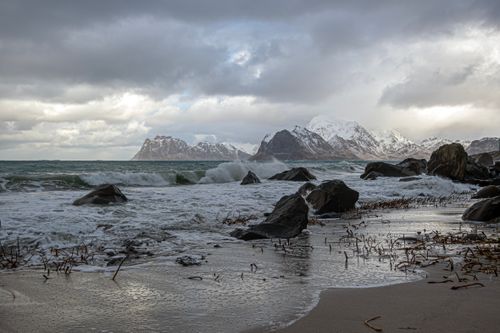 Free Waves crashing onto Myrland beach with snow-covered mountains in the background under moody skies. Stock Photo