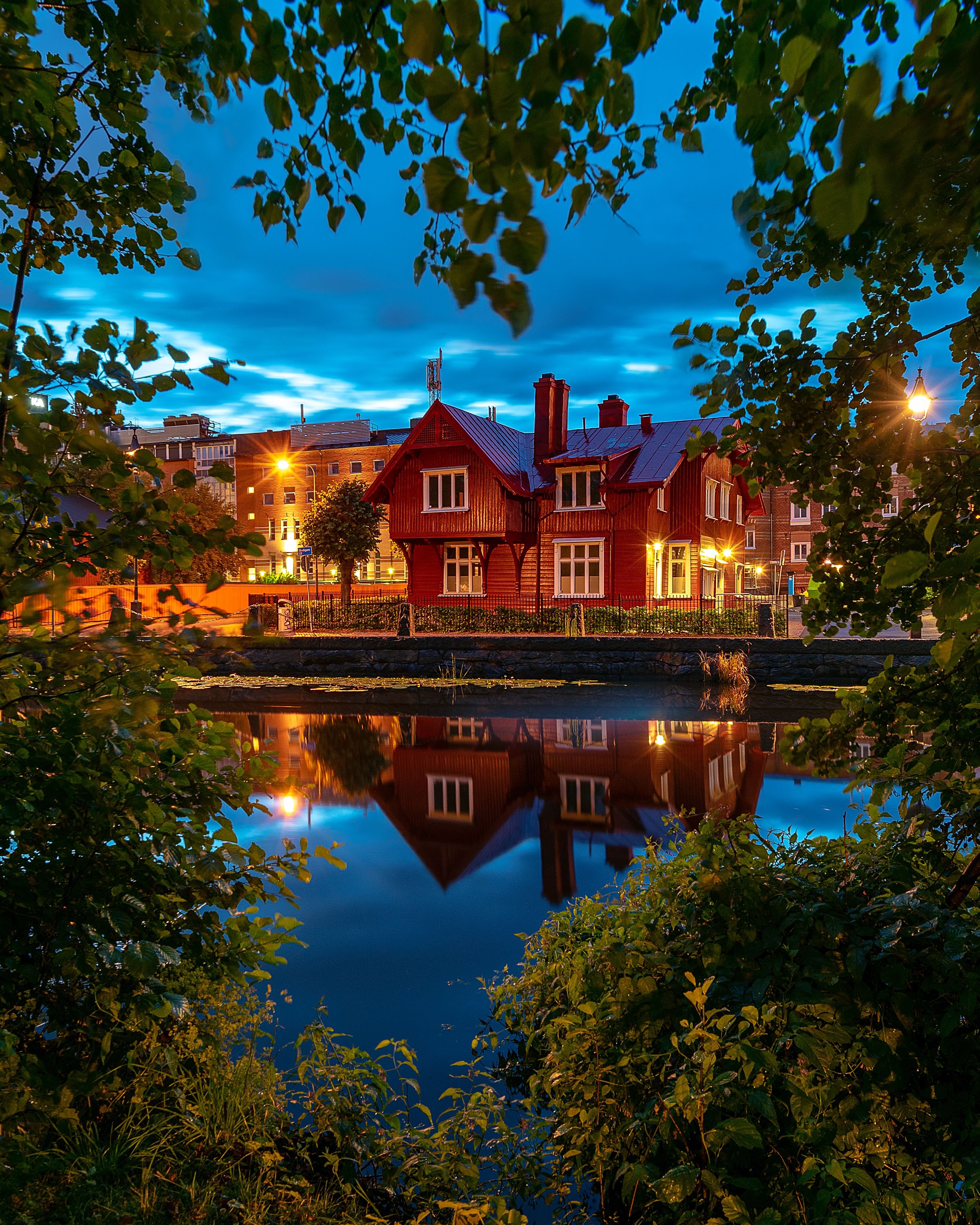 A picturesque view of a red house reflecting in the water, framed by trees in Göteborg, Sweden during twilight.