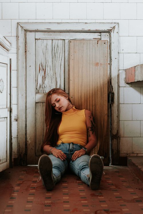 Free A young woman in casual attire sits relaxed against a vintage wooden door indoors, exuding a sense of calm. Stock Photo