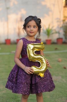 Cute little girl in a purple dress holding a gold number 5 balloon outdoors.