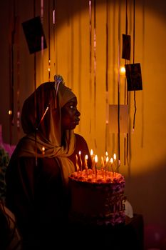 A woman in a hijab holds a birthday cake surrounded by candles in moody lighting.