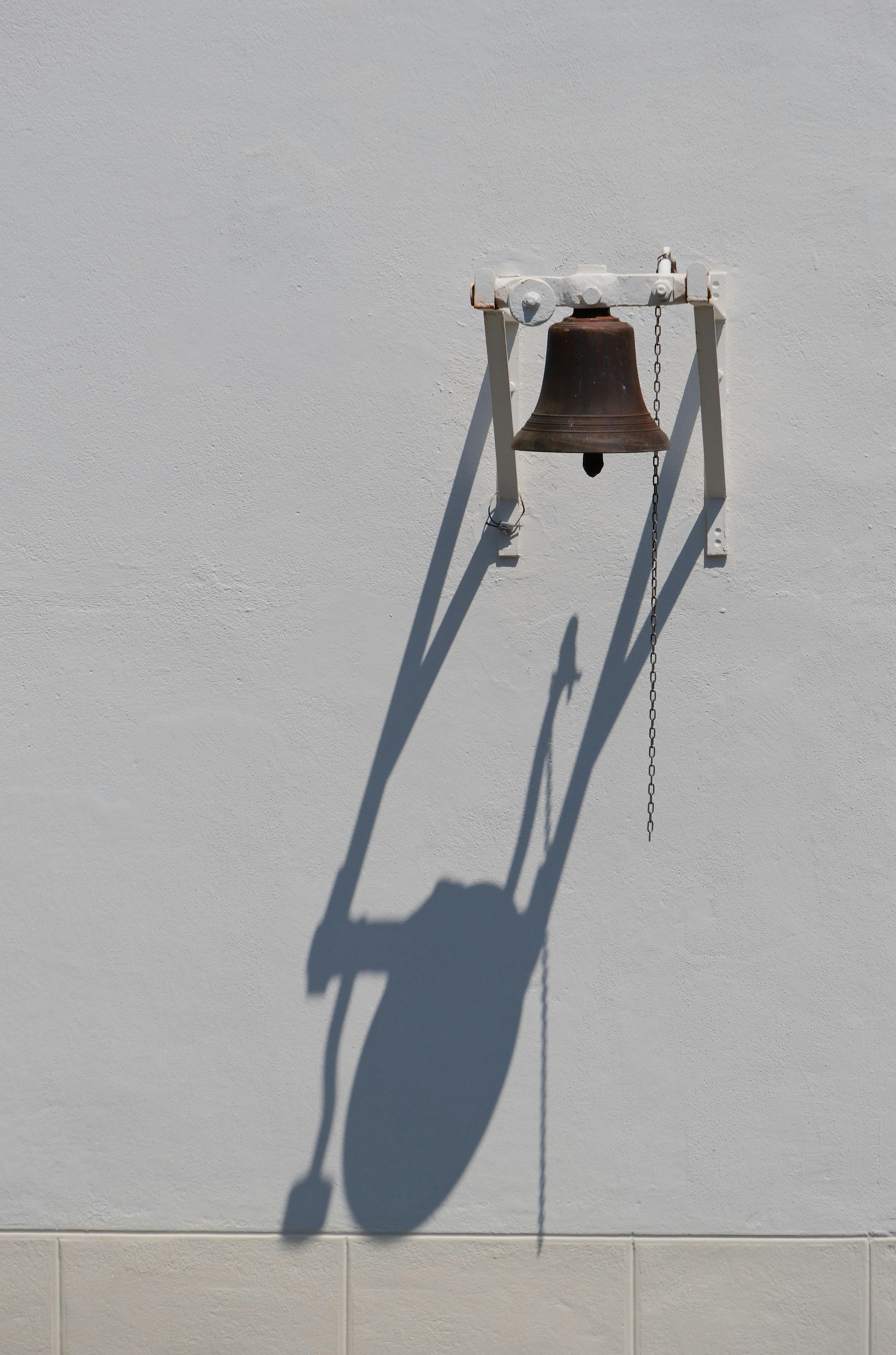 Ücretsiz Bronz bir çan, parlak güneş ışığı altında belirgin bir gölge oluşturarak beyaz bir duvara asılı duruyor ve minimalist tasarımı vurguluyor. Stok Fotoğraflar