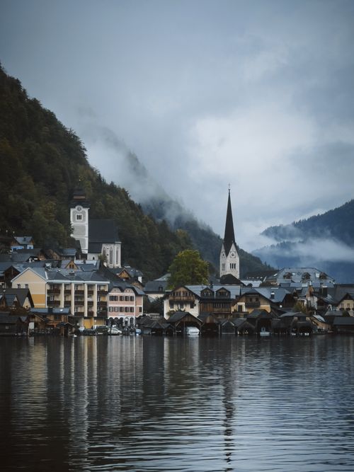 Free Misty morning over a charming lakeside village with iconic church spire and mountain backdrop. Stock Photo