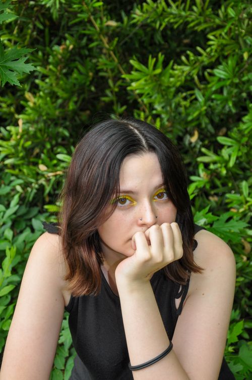 Free Portrait of a young woman with brown hair and yellow eyeshadow, resting her chin on her hand in a garden setting. Stock Photo