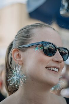 Joyful woman wearing sunglasses and glittering earrings enjoys a sunny outdoor event.