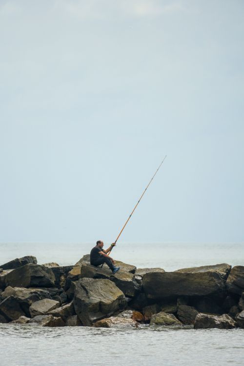 Free Solitary angler fishing on rocks by the sea under a cloudy sky, showcasing patience and natural serenity. Stock Photo
