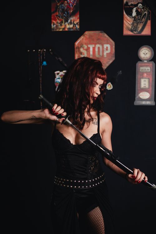 Free A woman in gothic attire holds a sword in a room with biker-themed decor, exuding a cool, edgy vibe. Stock Photo